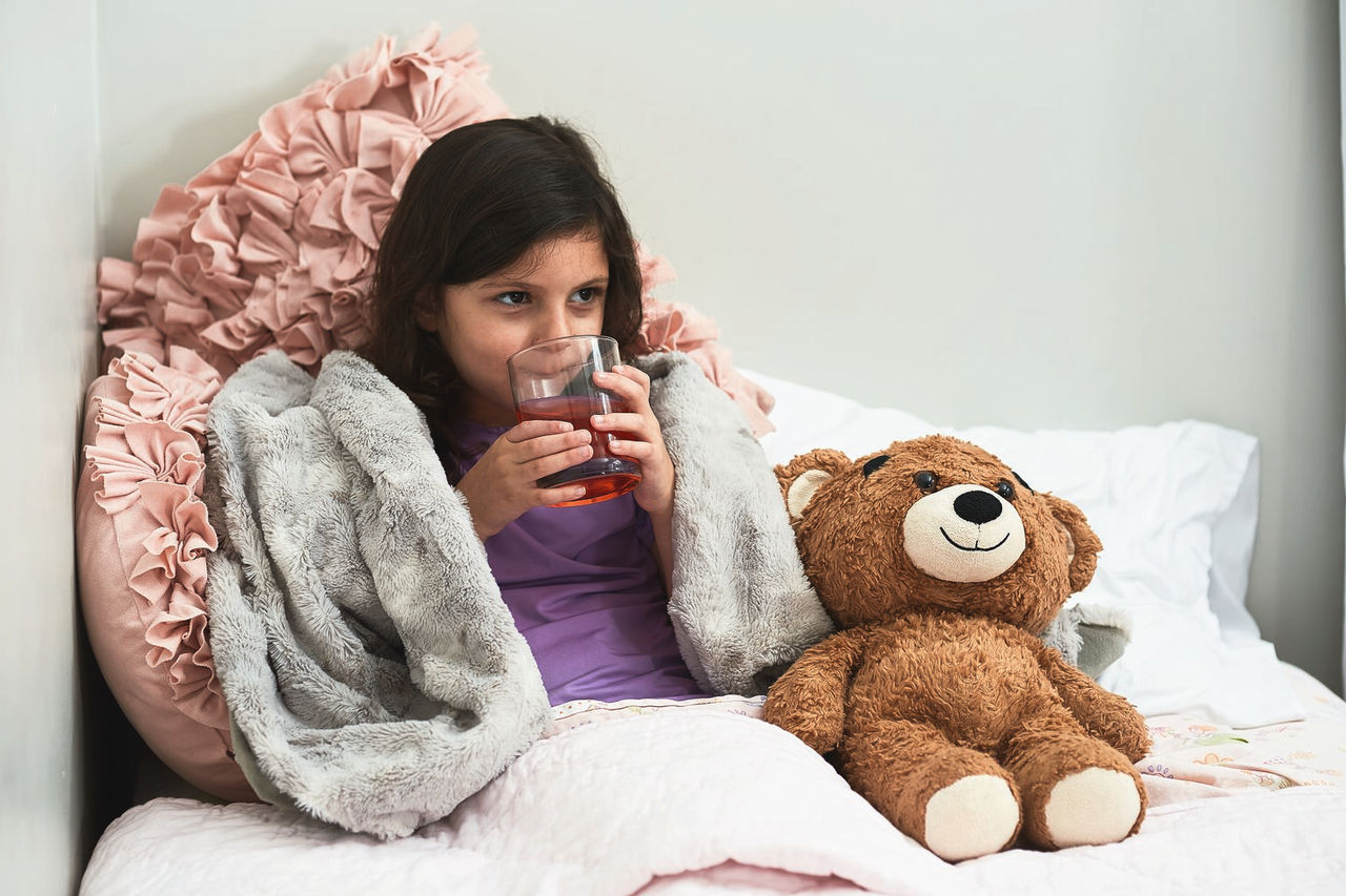 Dehydrated little girl sitting on her bed wrapped in a grey blanket surrounded by pink pillows and her teddy bear drinking a glass of liquid